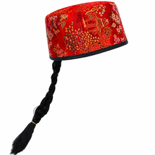 A red embroidered traditional cap with intricate gold and orange floral patterns, featuring a long black braided hair hanging from the side, set against a plain white background.