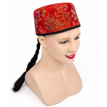 A red embroidered skullcap with a black band and long black braided hair, displayed on a beige mannequin head against a white background.