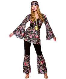 A woman modeling a 1960s-style hippie costume consisting of a black long-sleeve top with a colorful floral print, black pants, matching floral bell-bottom leg coverings, a black belt with a gold peace sign, and a matching headband, against a plain white background.
