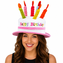 A woman wearing a novelty birthday hat shaped like a white cake with pink trim and six colorful candle toppers, against a plain white background.