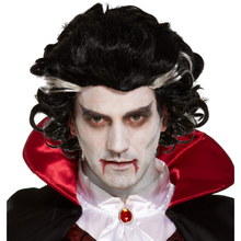 Man wearing a black vampire-style wig with dramatic curls, pale face makeup, red blood details on the lips and cheek, and a red-and-black cape with a white collar.