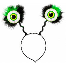 A novelty black headband with two large, furry, eyeball-like side decorations in green, white, and black, shown on a plain white background.