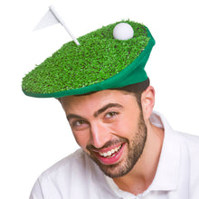 A man wearing a novelty golf hat shaped like a green putting surface with artificial grass, a small white flag, and a white golf ball on top, against a plain white background.