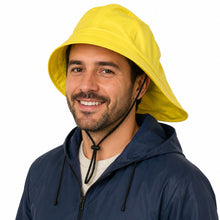 A smiling man wearing a bright yellow rain hat with a chin strap, paired with a navy blue hooded jacket, against a solid white background.