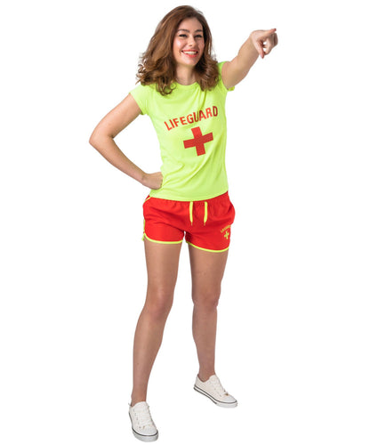 A woman modeling a bright lime green lifeguard t-shirt with a red cross and 'LIFEGUARD' text, paired with red swim shorts featuring yellow trim and white sneakers, set against a plain white background.