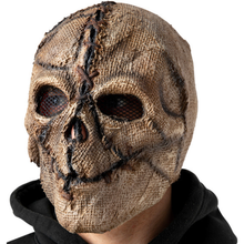 A skull-shaped burlap mask with distressed brown and black details, worn by a person in a black hoodie against a plain white background.