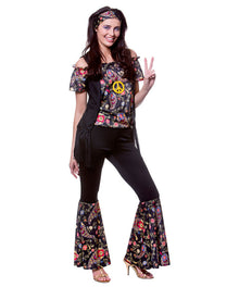 A woman modeling a 1960s-style hippie costume set against a plain white background. The set includes a black off-the-shoulder top with a colorful floral paisley print and a yellow peace sign on the chest, black bell-bottom pants with matching floral paisley print at the flared cuffs, a matching floral headband, and metallic high heels.