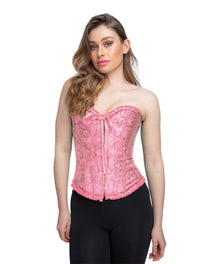 A woman modeling a pink lace corset top with a sweetheart neckline, center front tie, and ruffled trim, paired with black pants against a plain white background.