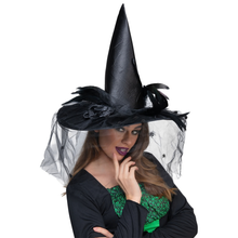A woman wearing a tall black witch hat with black feathers, sheer black tulle, and spiderweb details, paired with a black top and green lace underlayer, against a plain white background.